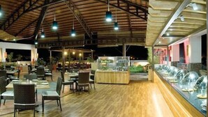 A spacious indoor dining area with multiple tables and chairs, a central serving station, and a glass display case on the right. - Ramyas Hotels (Tiruchirappalli)