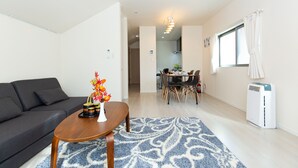 Ein modernes Wohnzimmer mit einem dunklen Sofa, einem Holztisch und einem gemusterten Teppich. Im Hintergrund ist ein Essbereich mit Tisch und Stühlen zu sehen.