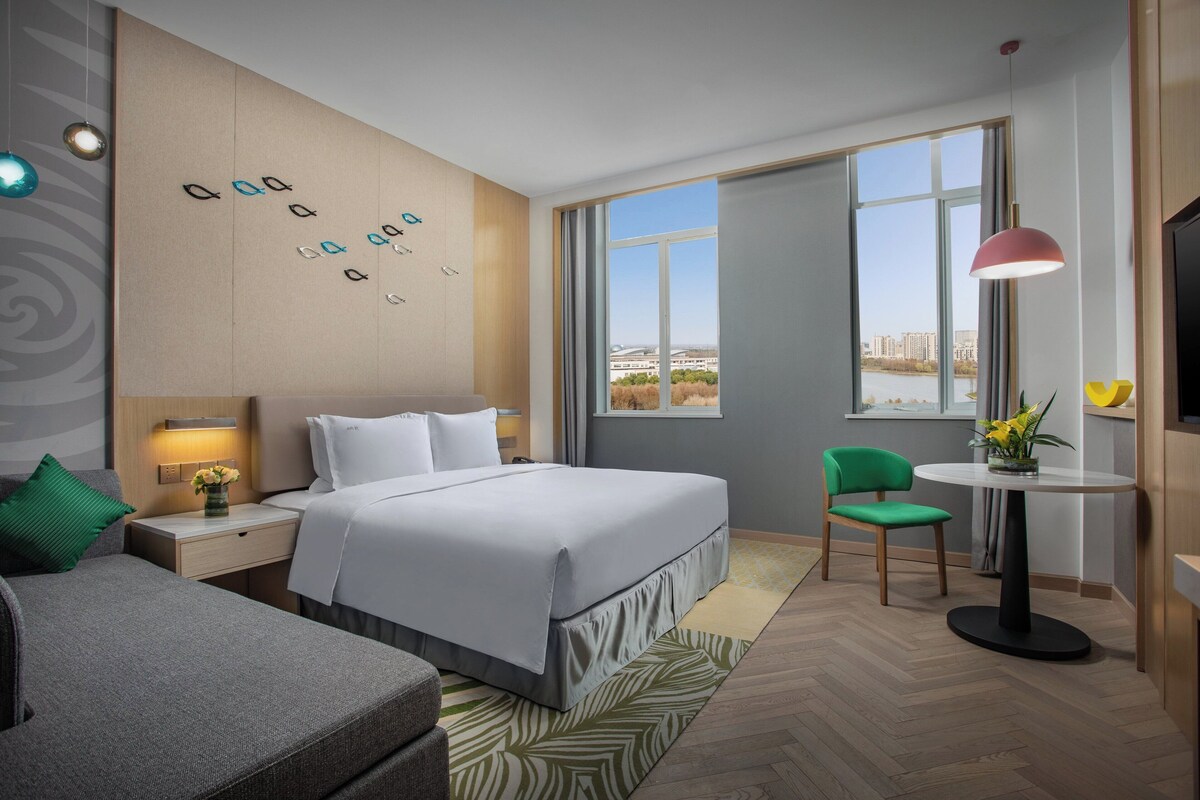A modern hotel room with a large bed, a desk, a chair, and a view of the city.