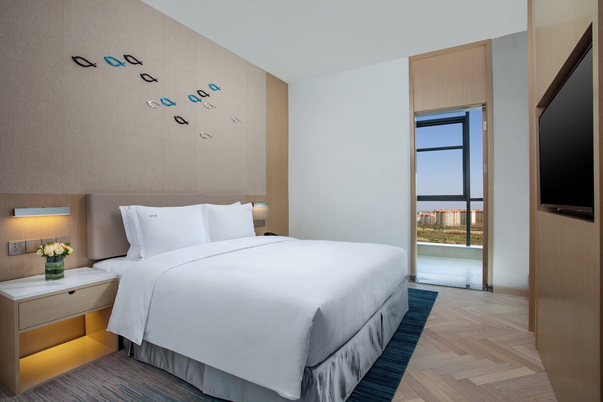 A modern hotel room with a large bed, a nightstand with a vase of flowers, a wall-mounted light, and a large window offering a city view.