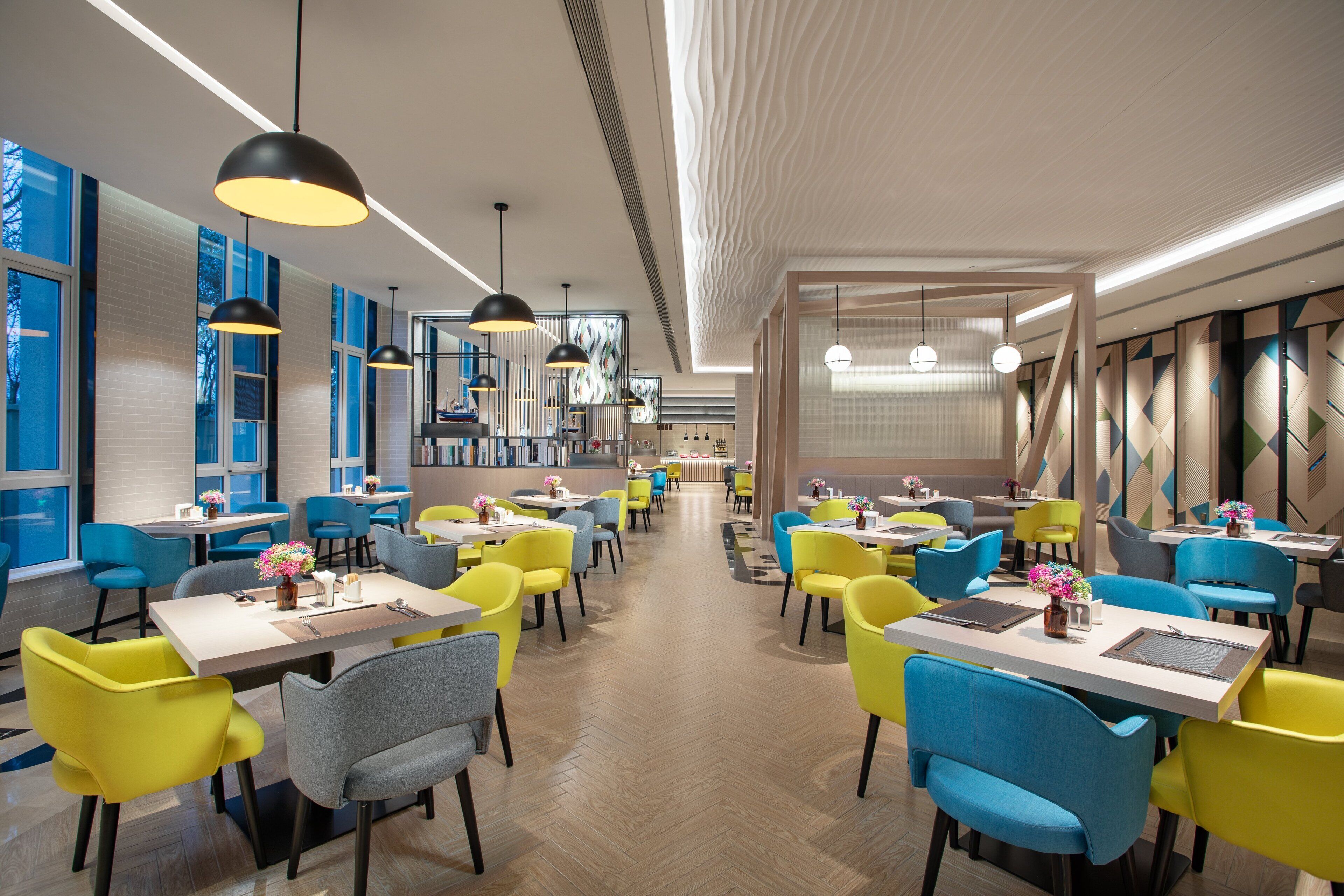 Un restaurant moderne avec des chaises jaunes et bleues, des suspensions noires et un motif de parquet en chevrons.