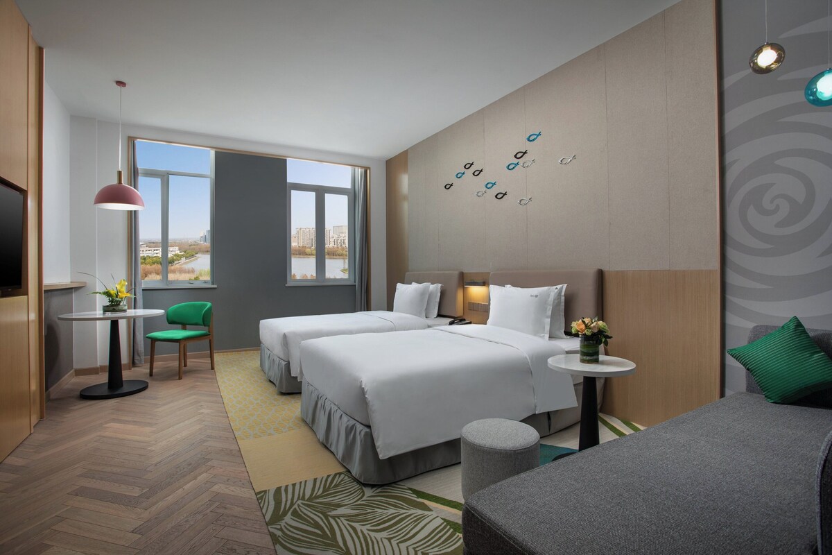 A hotel room with two beds, a small table, a chair, a TV, and a view of the city.