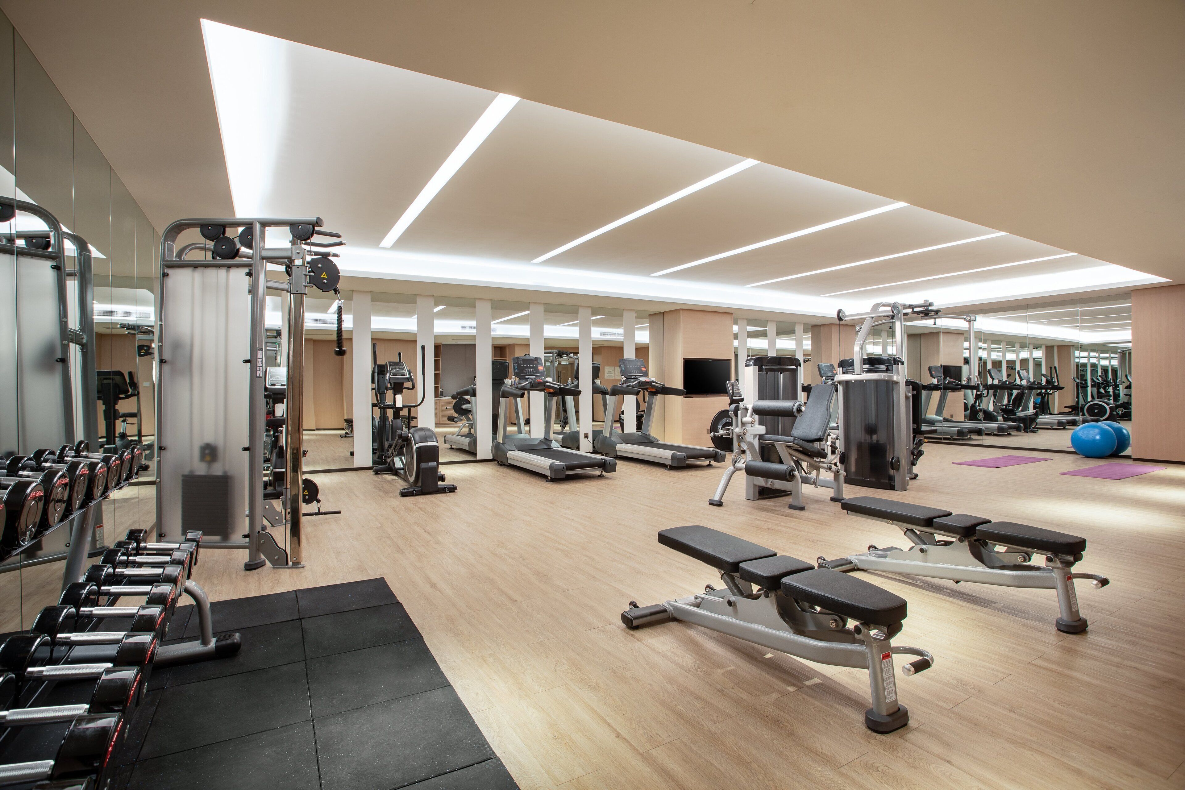 A modern gym with various exercise equipment, mirrored walls, and a wooden floor.