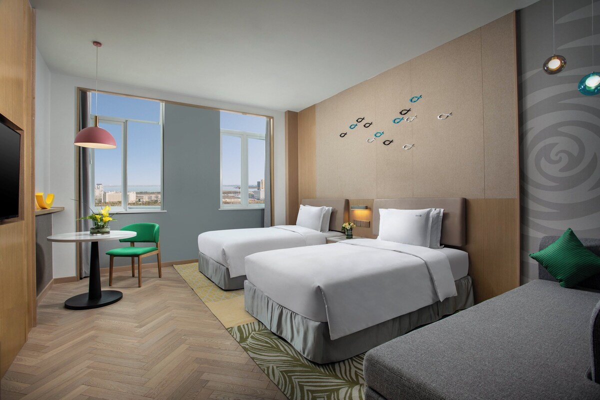 A hotel room with two beds, a dining area with a table and chairs, a TV, and a view of the city.