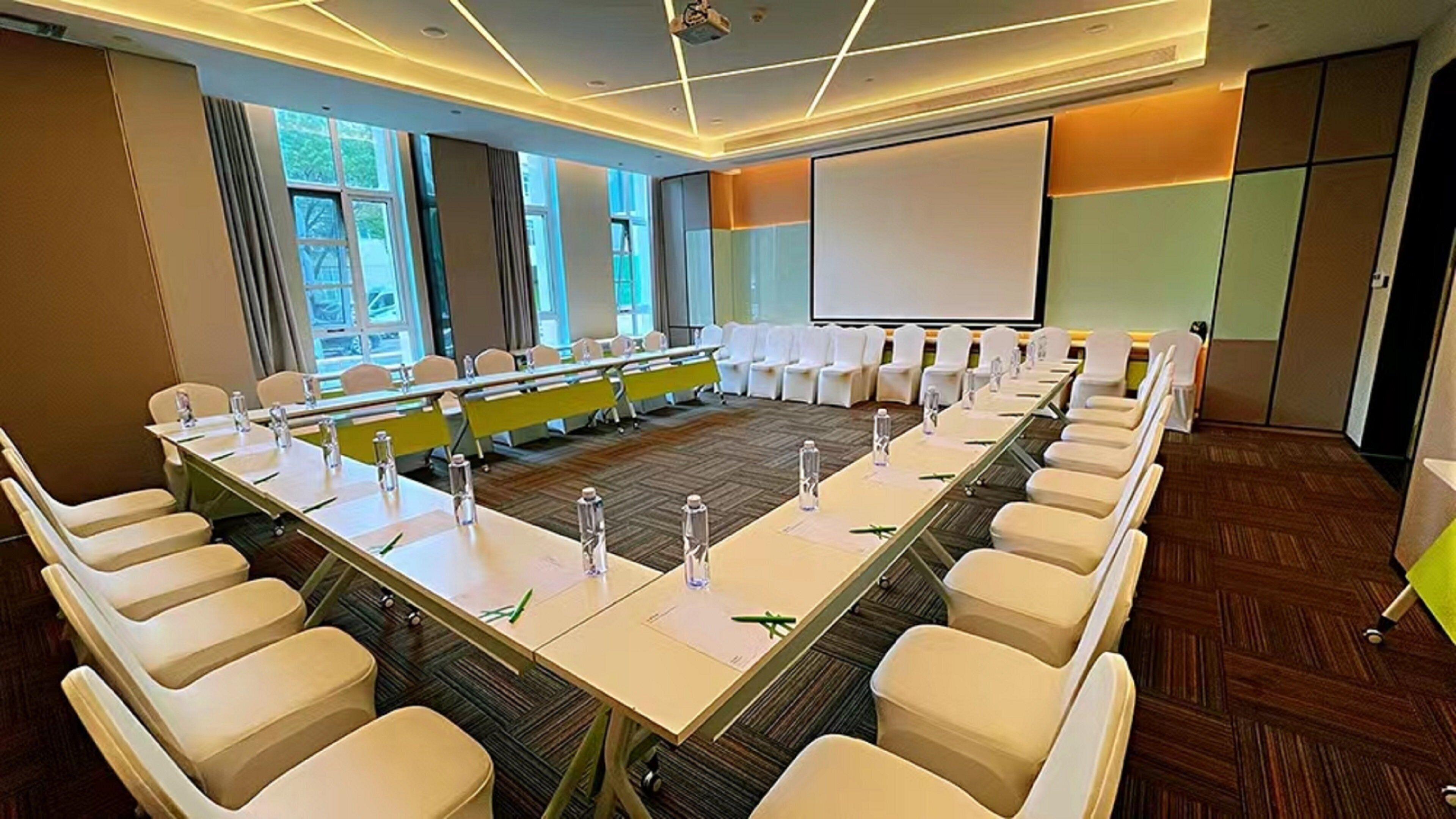 A conference room with a long table, white chairs, and a projector screen.