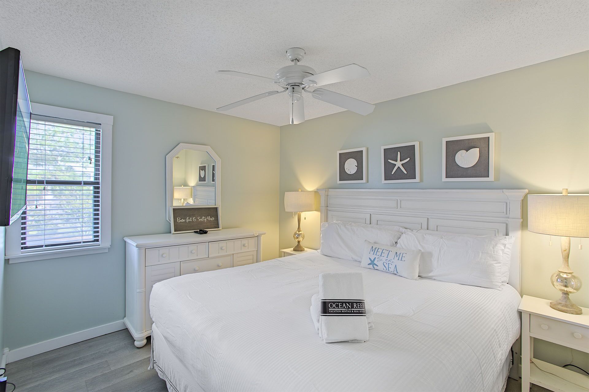 A bedroom with a bed, a ceiling fan, a dresser, a nightstand, and wall art.