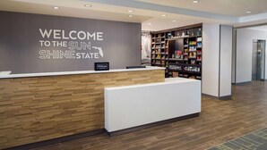 A hotel reception desk with a 'Welcome to the Sun Shine State' sign, a reception area, and a bookshelf with various items. - Hampton Inn Kissimmee North (Kissimmee)