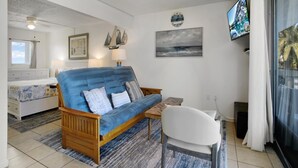 Uma sala de estar moderna com um sofá azul, mesa lateral de madeira e uma cadeira branca. Há uma janela grande com cortinas, um ventilador de teto e uma TV fixada na parede.
