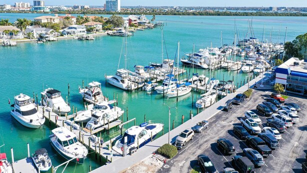 Marina com vários barcos atracados, área de estacionamento com carros e vista para o horizonte urbano.