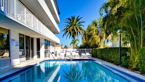 Piscina com espreguiçadeiras, uma palmeira e um edifício com varanda.