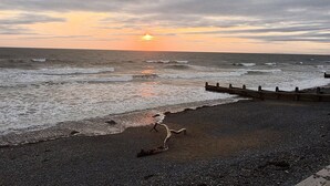 A beach with a wooden walkway leading to the water, waves crashing onto the shore, and a sunset in the background. - Angorfa Family Holiday Home with Games Room (Tywyn)