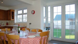 A dining area with a table set for six, a clock on the wall, and large windows with a view of a house and lawn. - Angorfa Family Holiday Home with Games Room (Tywyn)
