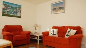 A living room with a red armchair, a red sofa with anchor pillows, a small white table with a lamp, and a painting of a coastal scene. - Angorfa Family Holiday Home with Games Room (Tywyn)
