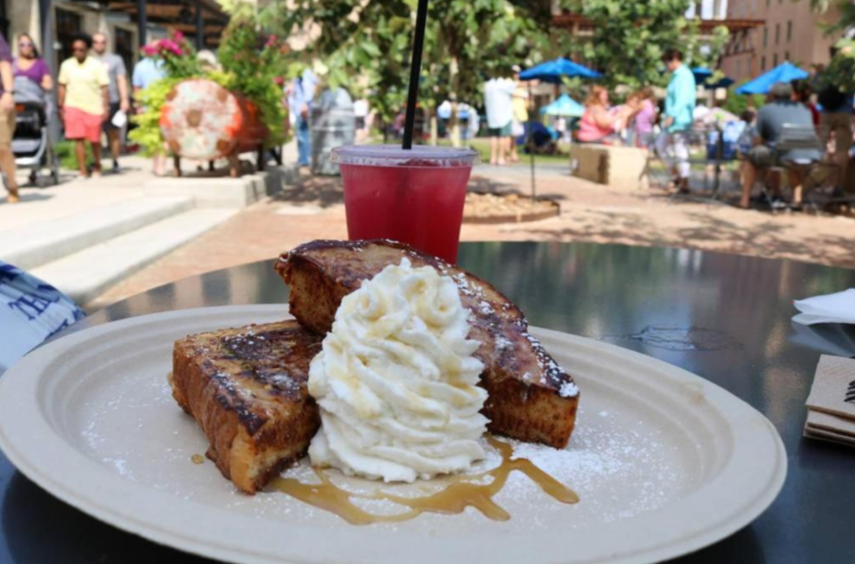 Ein Teller French Toast mit Schlagsahne und Sirup, ein rotes Getränk im Hintergrund und eine ungezwungene Essgelegenheit im Freien.