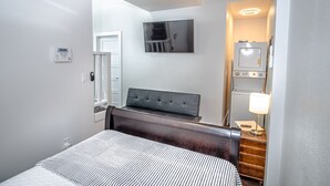 Ein modernes Hotelzimmer mit einem Bett, einem an der Wand befestigten Fernseher, einem Nachttisch mit Lampe und einem kleinen Schreibtisch mit Computer.
