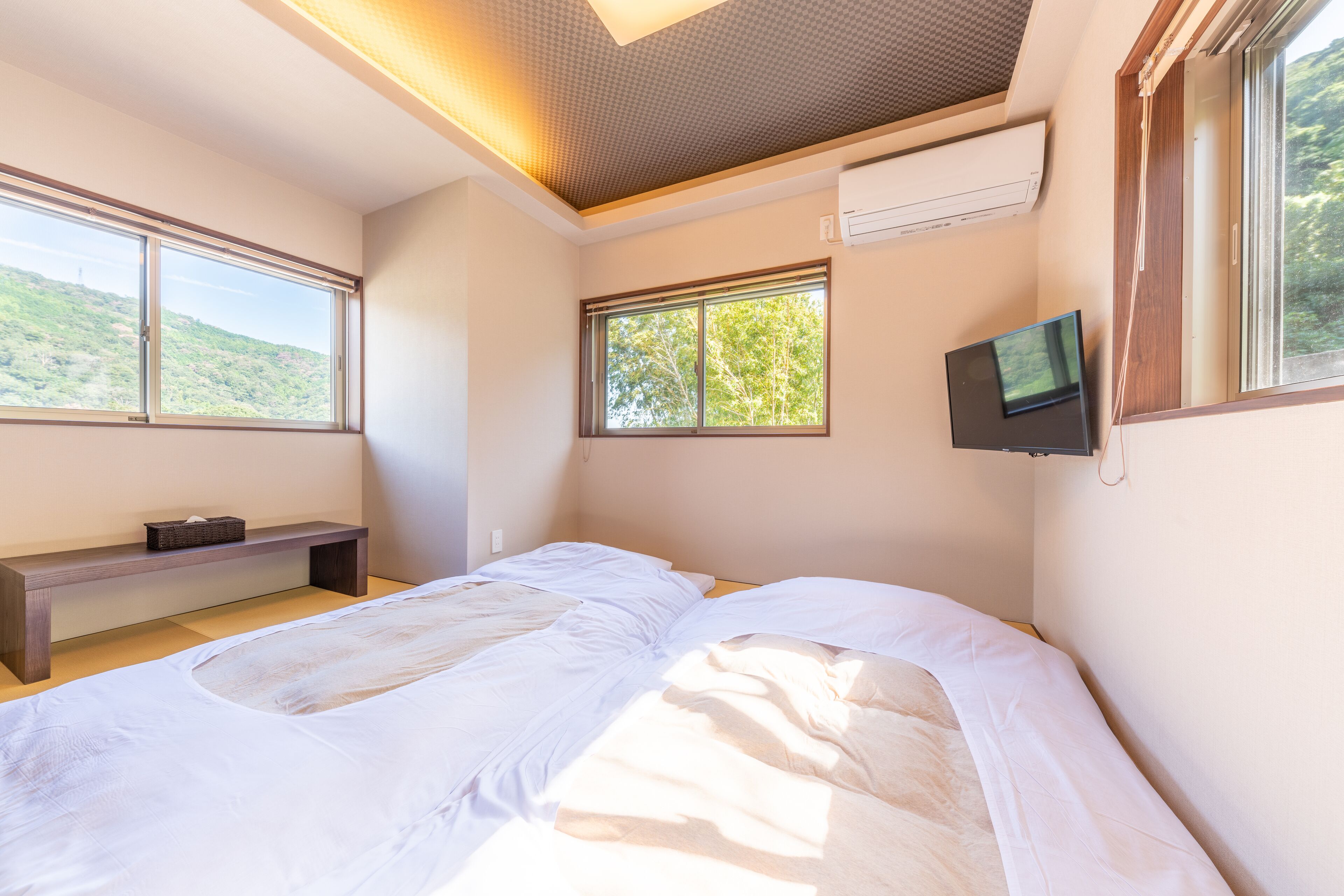 A bedroom with a large bed, a bench, and two windows offering views of a green landscape.