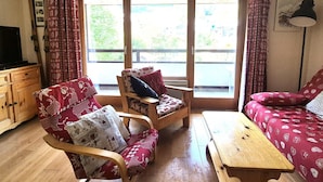 Un salon chaleureux avec un canapé, deux fauteuils, une table basse en bois et un téléviseur posé sur un meuble en bois. De grandes fenêtres, ornées de rideaux, laissent entrer la lumière naturelle et offrent une vue sur l’extérieur.