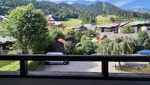 Une vue depuis un balcon donnant sur un village avec des bâtiments traditionnels, une route et une chaîne de montagnes boisée.