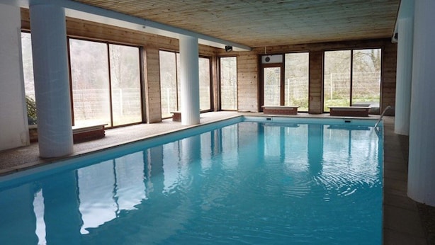 Une piscine intérieure avec de grandes baies vitrées, des poutres en bois et un banc le long du bord.