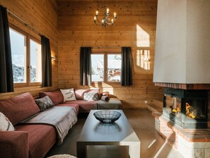 Een gezellig blokhutinterieur met een bank, een tafel met een kom, een open haard en grote ramen.