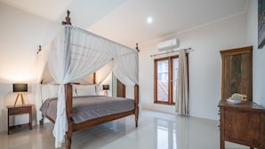 Una cama con dosel sobre cuatro postes, con estructura de madera y cortinas blancas, una mesita de noche con lámpara y un mueble de madera.