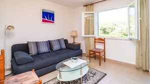 Uma sala de estar com um sofá azul escuro, uma mesa de centro de vidro, uma cadeira de madeira, um abajur e uma janela com cortinas.