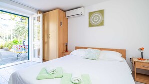 Quarto de hotel com cama, mesa de cabeceira, ar condicionado, janela com vista para o verde e porta que dá para o exterior.