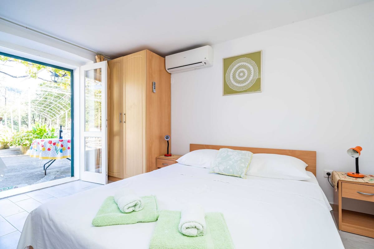 Une chambre d’hôtel comprenant un lit, une table de chevet, un climatiseur, une fenêtre donnant sur un espace verdoyant et une porte menant à l’extérieur.