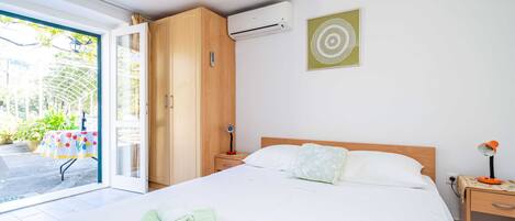 Une chambre d’hôtel comprenant un lit, une table de chevet, un climatiseur, une fenêtre donnant sur un espace verdoyant et une porte menant à l’extérieur.