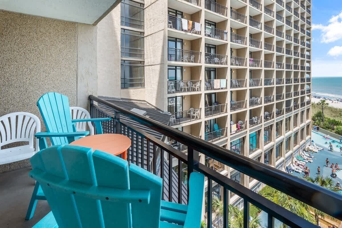 A balcony with turquoise chairs and a small table, overlooking a pool and a beach.