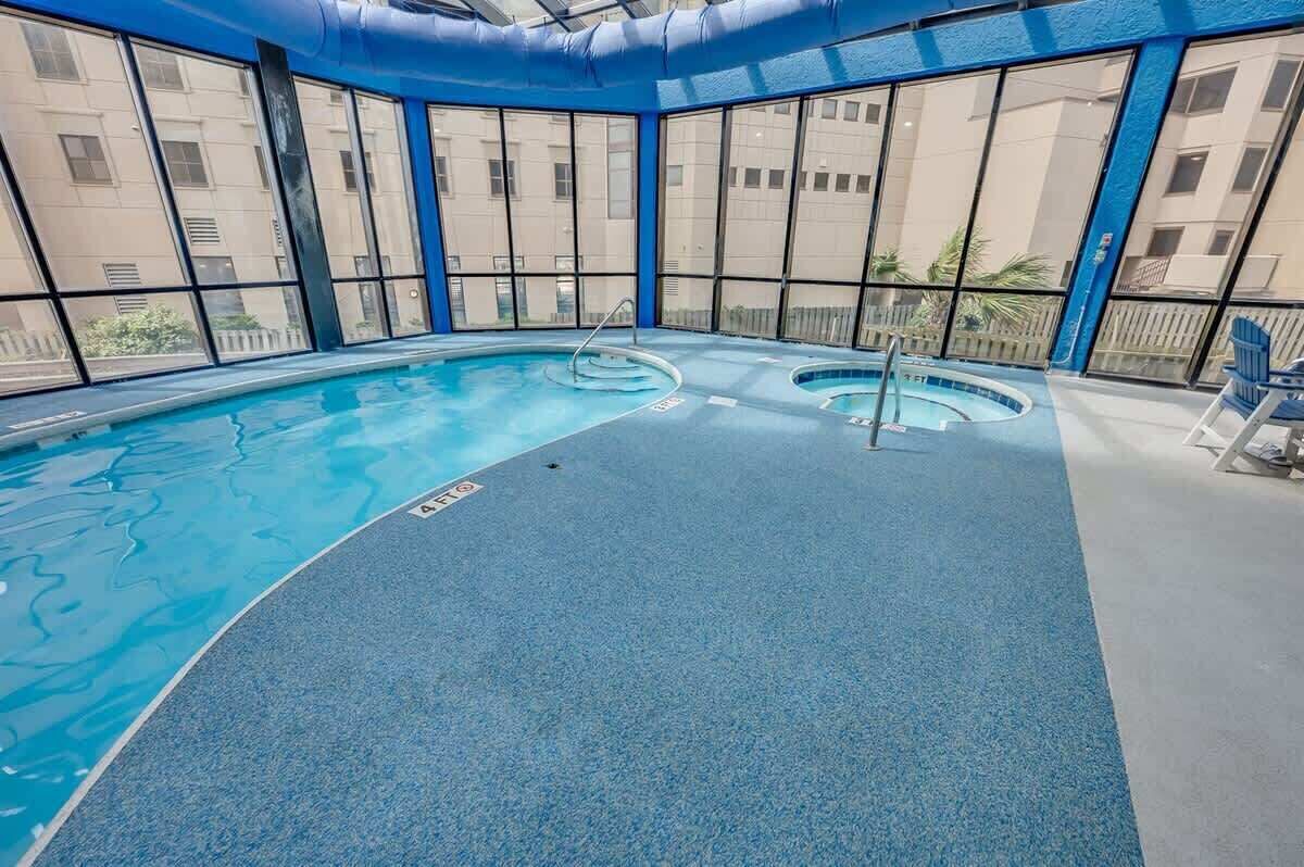 An indoor swimming pool with clear blue water, surrounded by large windows offering views of buildings outside.