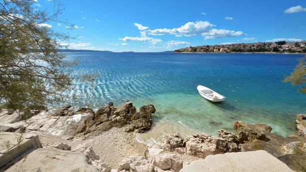 A coastal scene with clear blue water, a boat, rocky shore, and a distant town. - SOLEA - 2 bedroom sea view apartment 100m from the beach in Okrug Gornji (Okrug Gornji)