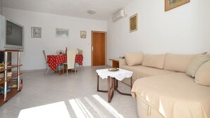 A living room with a beige sofa, a small table with a lace tablecloth, a television, and a dining area with a red and white checkered tablecloth. - SOLEA - 2 bedroom sea view apartment 100m from the beach in Okrug Gornji (Okrug Gornji)