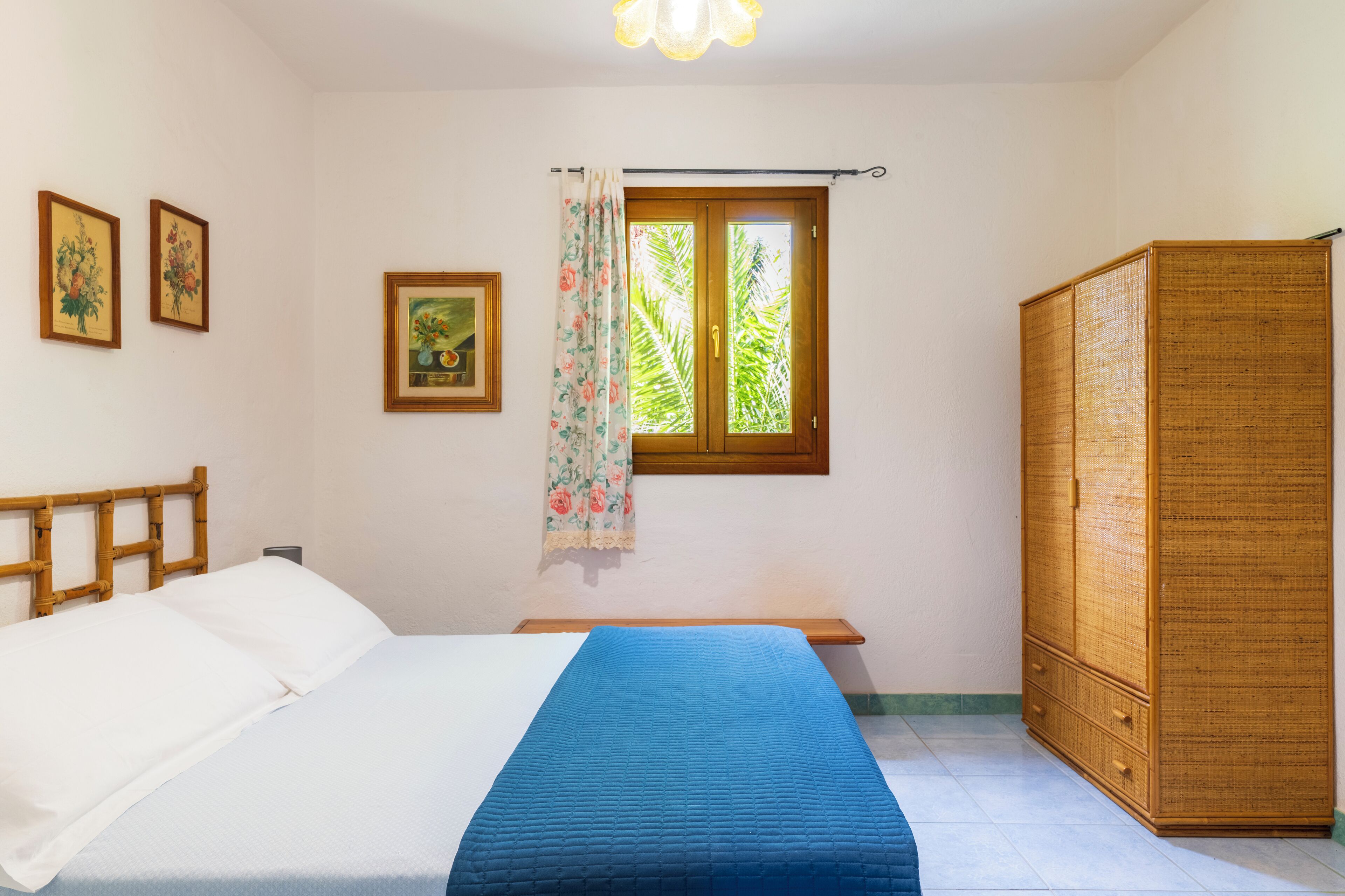A bedroom with a bed, a wardrobe, a window with curtains, and framed pictures on the wall.