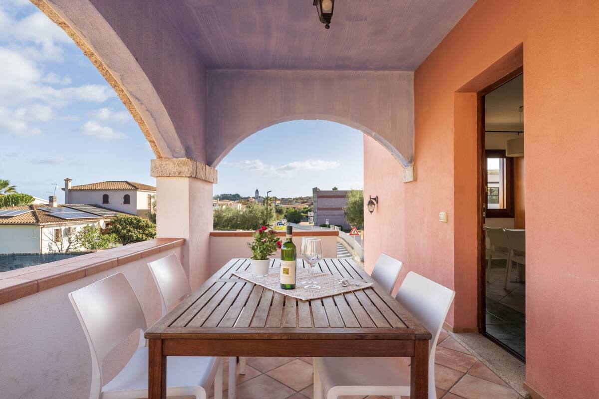 Um alpendre coberto com uma mesa de madeira, cadeiras brancas e uma garrafa de vinho.