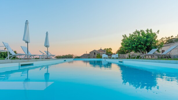 A swimming pool with lounge chairs and umbrellas, surrounded by stone buildings and greenery. - Baglio Cella, Basil annex in the Hyblean countryside (Modica)