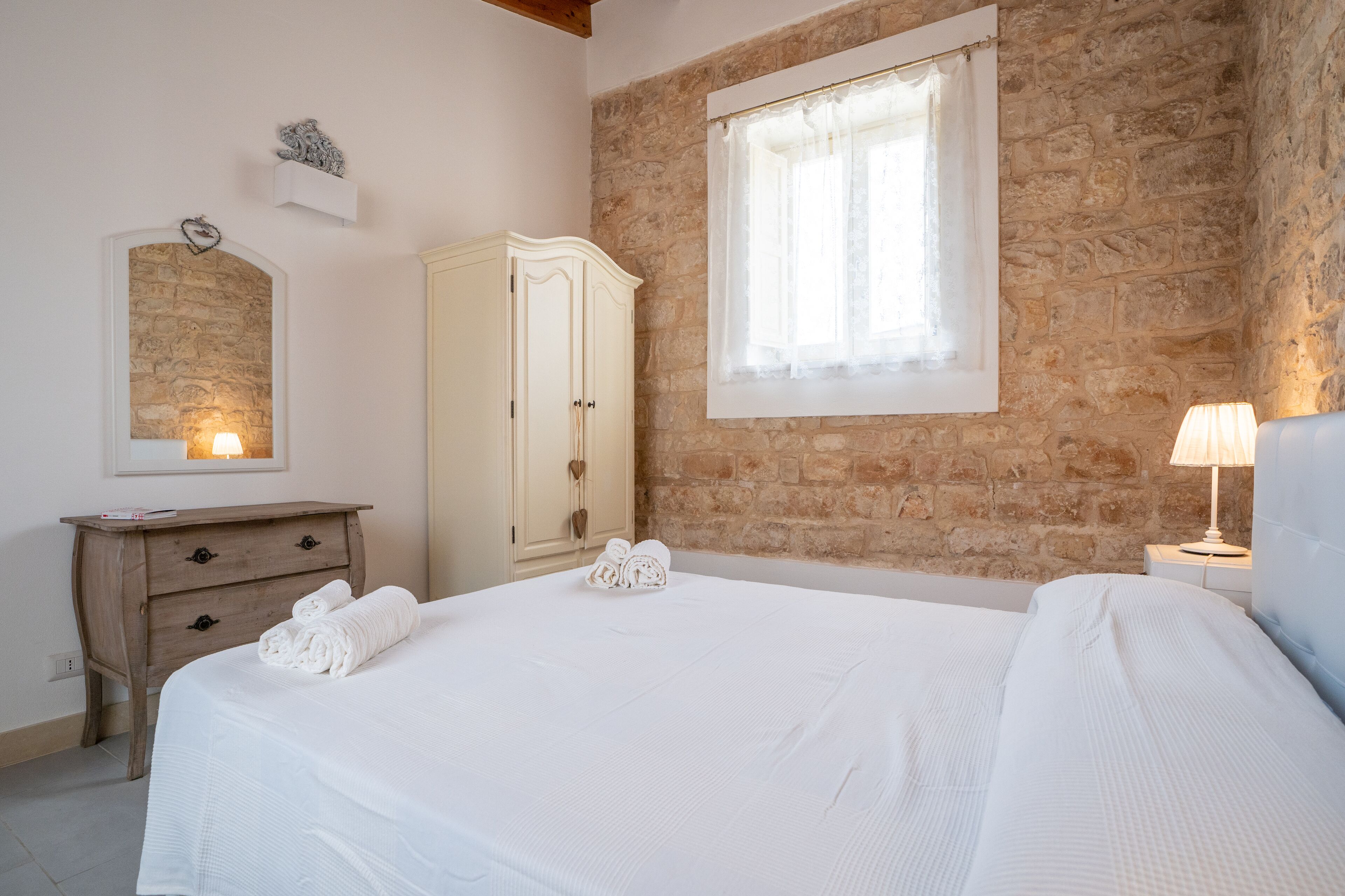 A bedroom with a stone wall, a wooden nightstand with a lamp, a white bed with rolled towels, a wooden dresser, and a mirror.