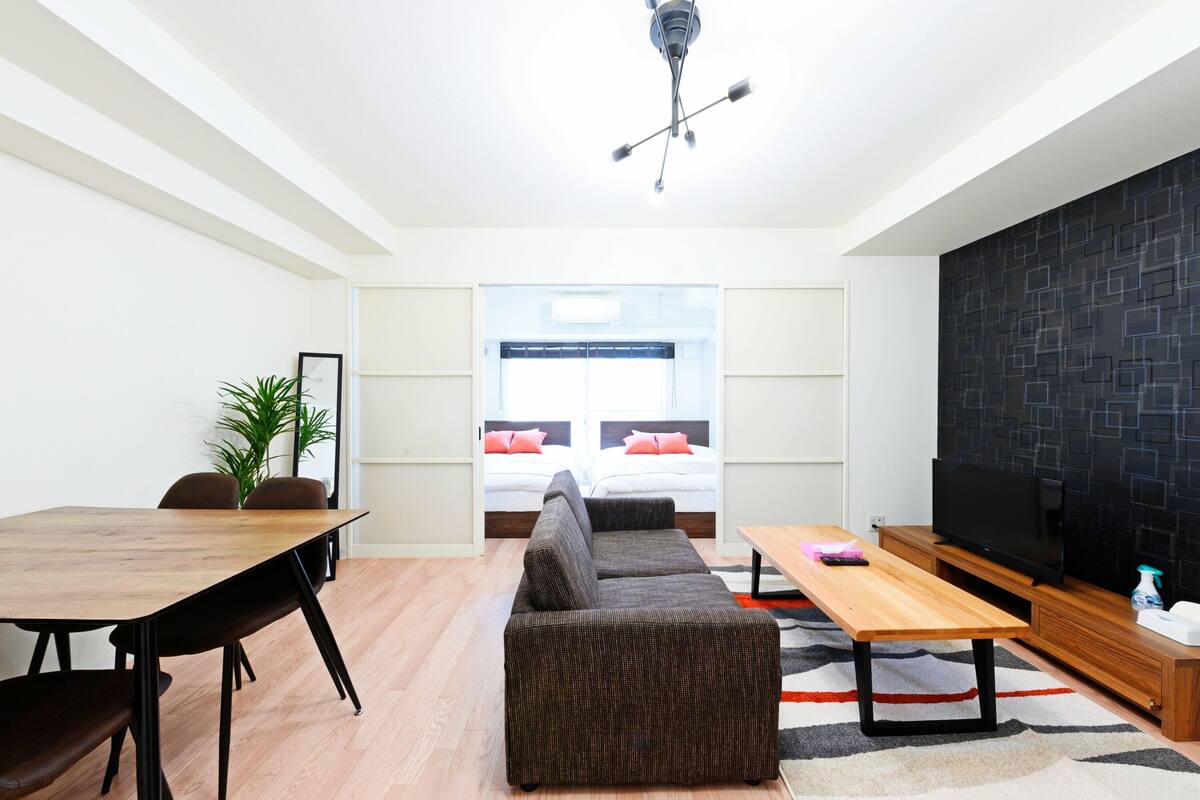 A modern living room with a sofa, a dining table, and a TV. There is a window with a view of a bed and a wall with a geometric pattern.