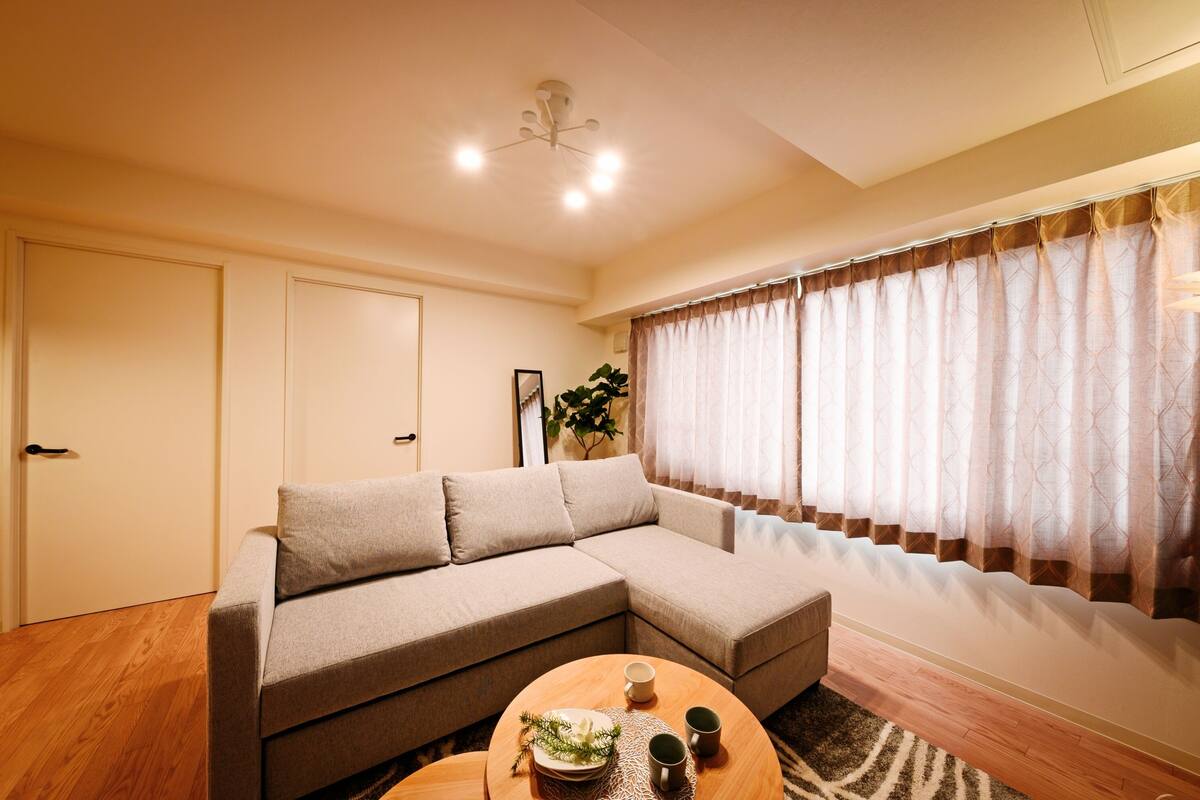 A living room with a beige sectional sofa, a wooden coffee table, and a large window with patterned curtains.