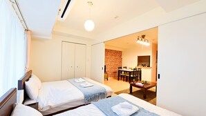 Une chambre d’hôtel avec deux lits, un coin repas avec un mur en briques et une cuisine moderne visible à travers une porte ouverte.