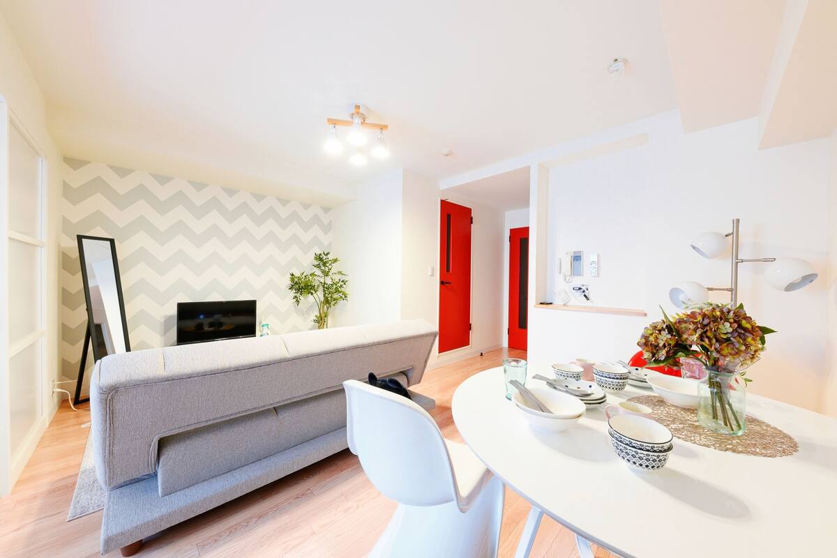 A modern living room with a sofa, a dining table with bowls and a vase, and a TV in the background.