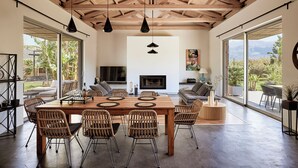 Un vaste espace de salle à manger avec une table en bois et des chaises en osier, de grandes fenêtres offrant une vue sur la verdure, et une cheminée en arrière-plan.
