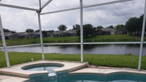 A swimming pool with a curved edge, surrounded by a covered area with a view of a lake and houses in the background. - Lake Therapy, Experience the ultimate vacation getaway in vibrant Caggans Crossing. (Clermont)