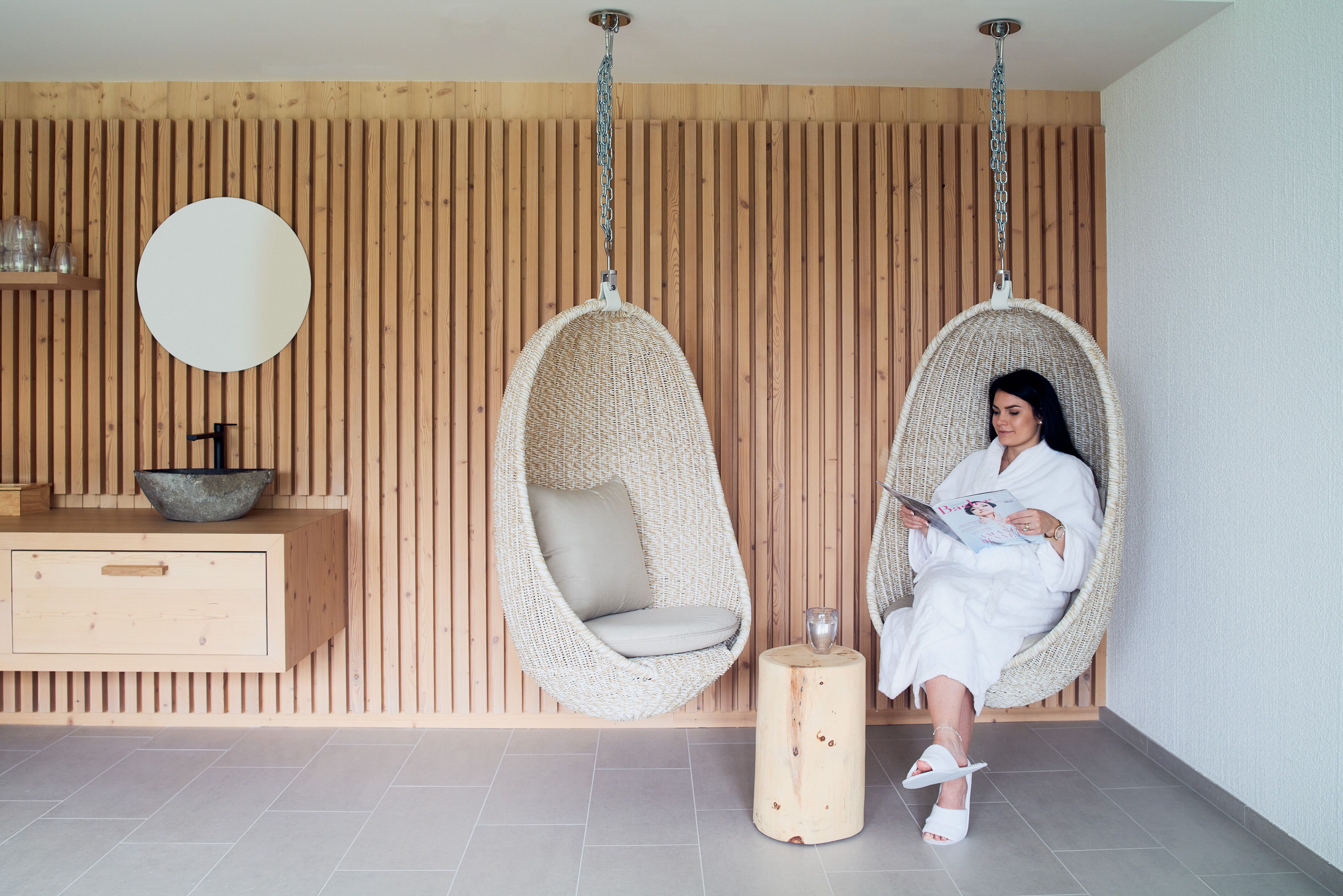 Una habitación con dos sillas colgantes, un muro de madera, un espejo redondo, un armario de madera y una pequeña mesa redonda.