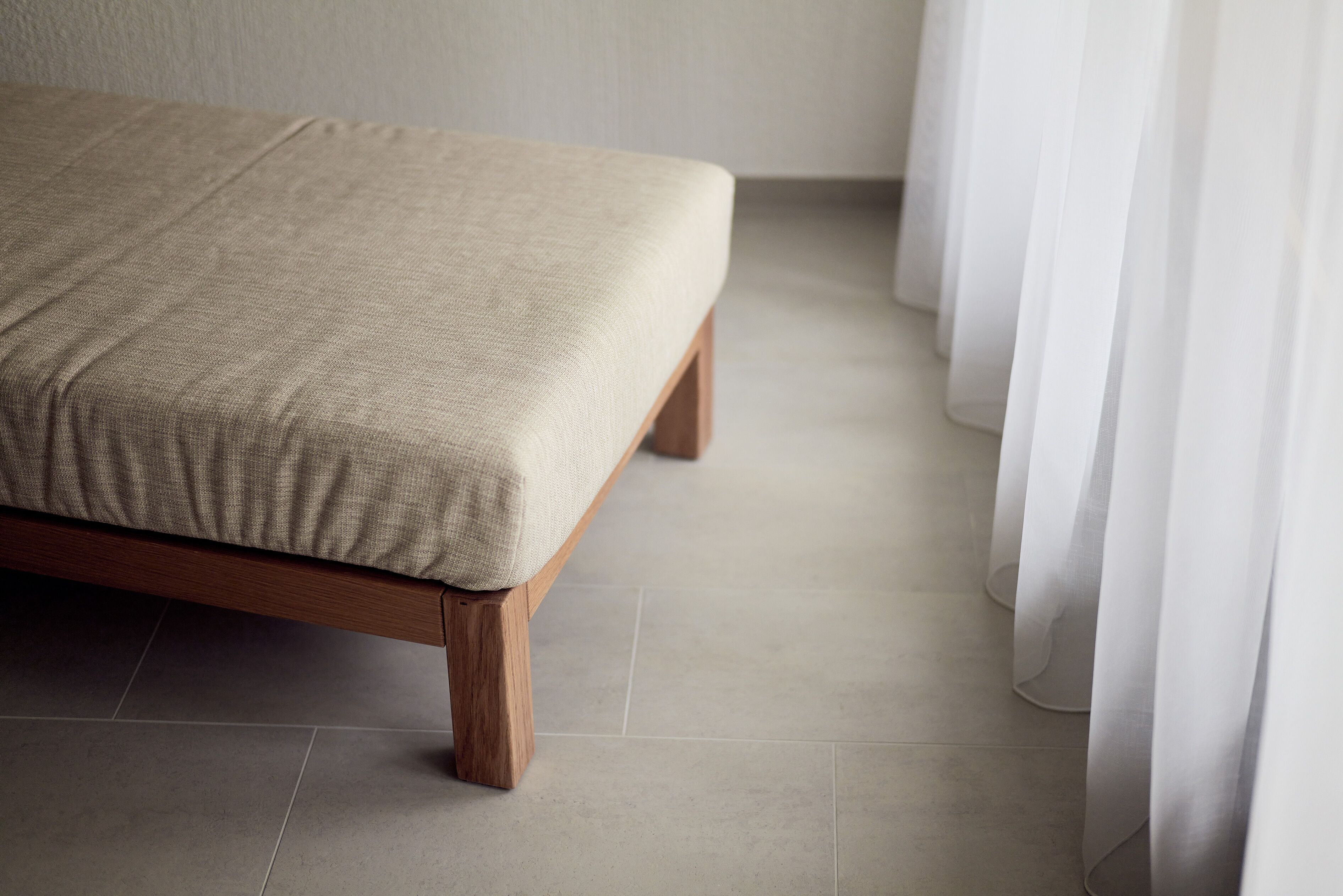 A bed with a beige bedspread and wooden legs, positioned against a wall with a curtain partially drawn to the side.