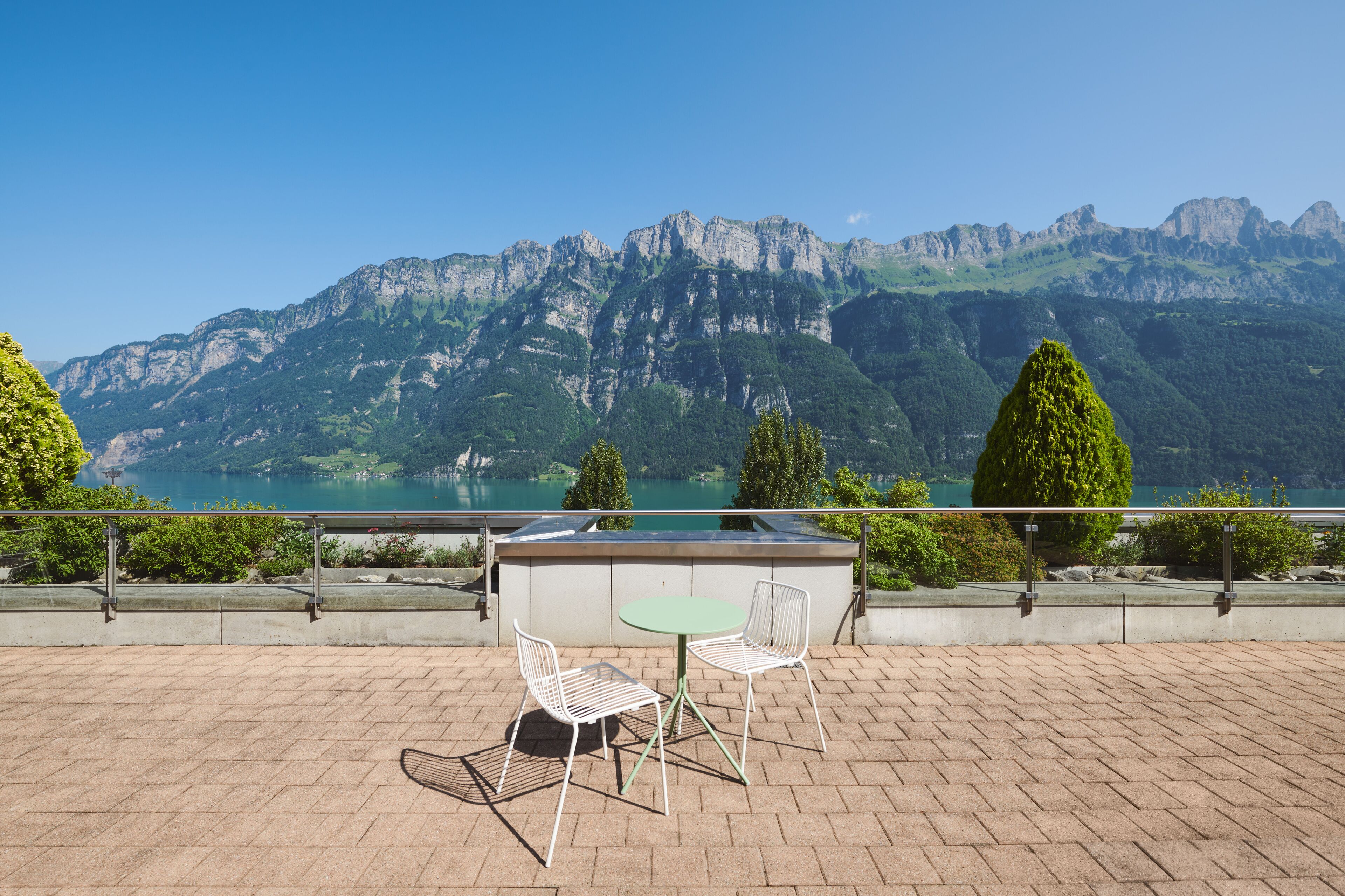 A terrace with two white metal chairs and a small round table, overlooking a mountainous landscape with a lake.