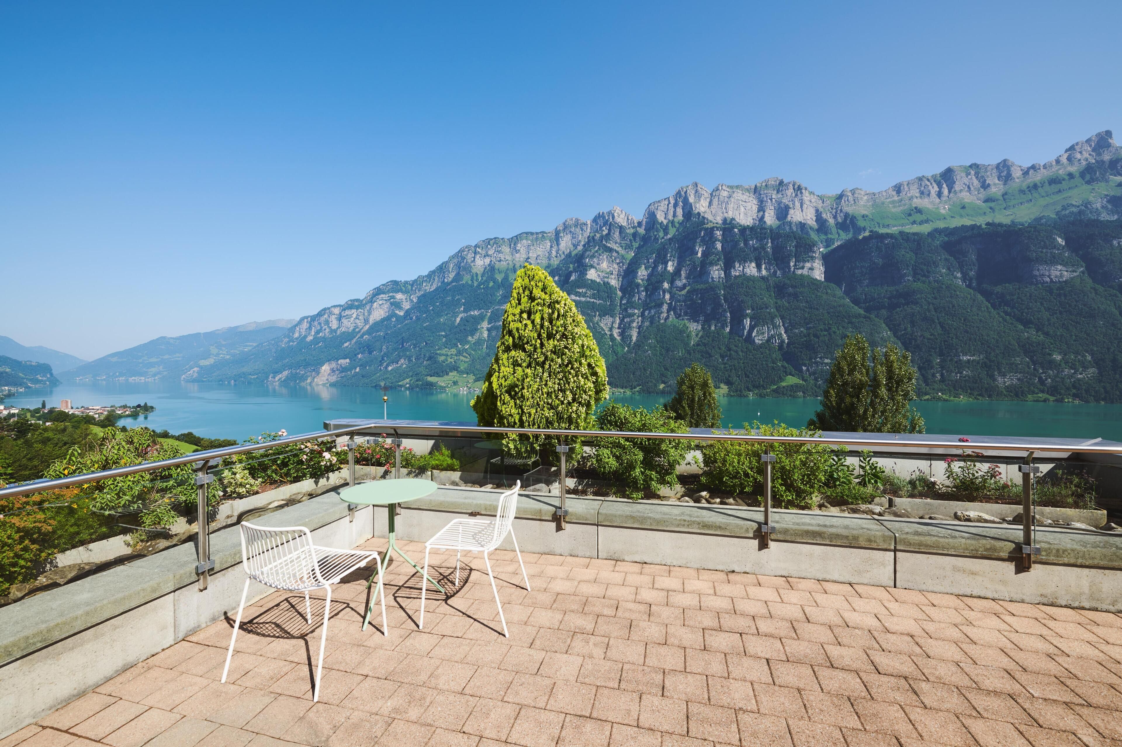 Una terraza con dos sillas blancas y una mesa, con vistas a un paisaje montañoso y un lago.
