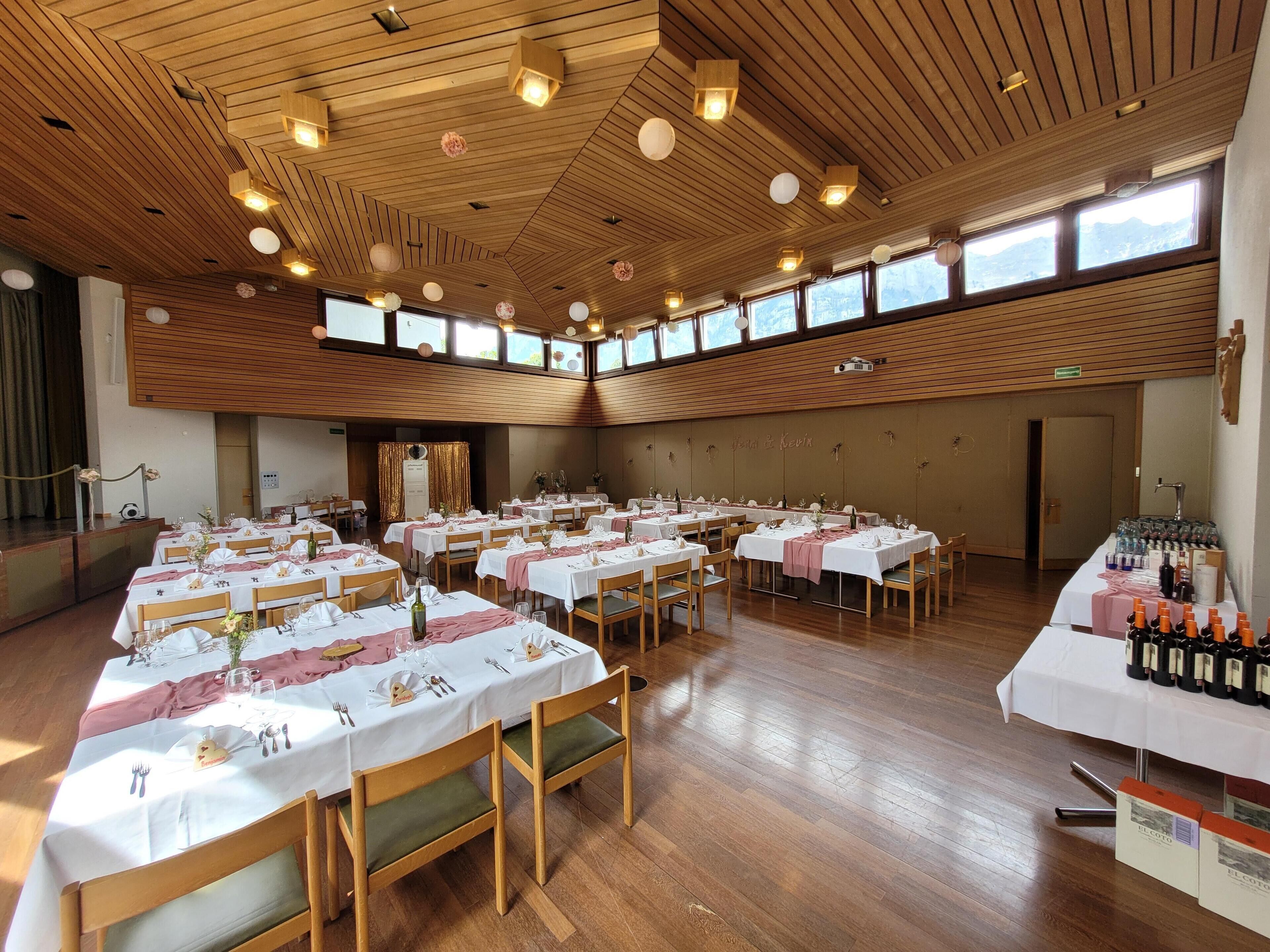 A spacious hall with multiple tables set for dining, wooden ceiling with recessed lighting, and large windows providing natural light.