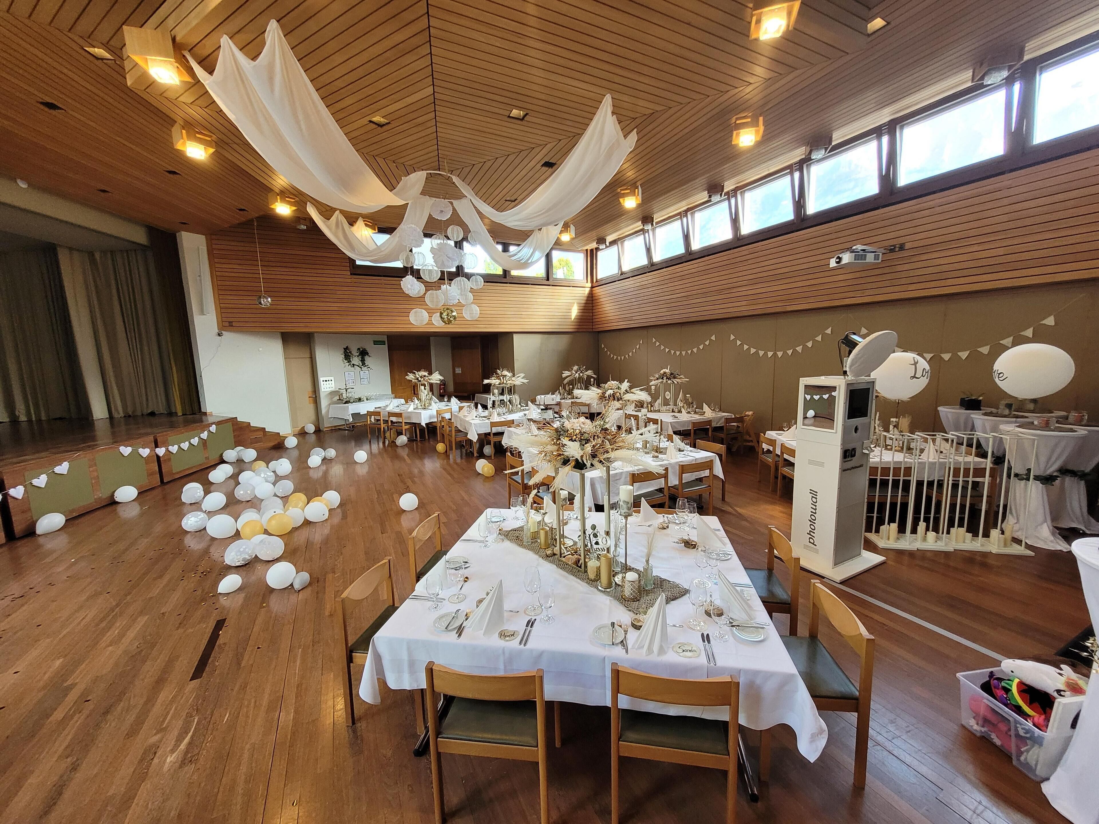 A spacious hall with wooden floors and high ceilings, featuring tables set for a formal event with white tablecloths and floral centerpieces.