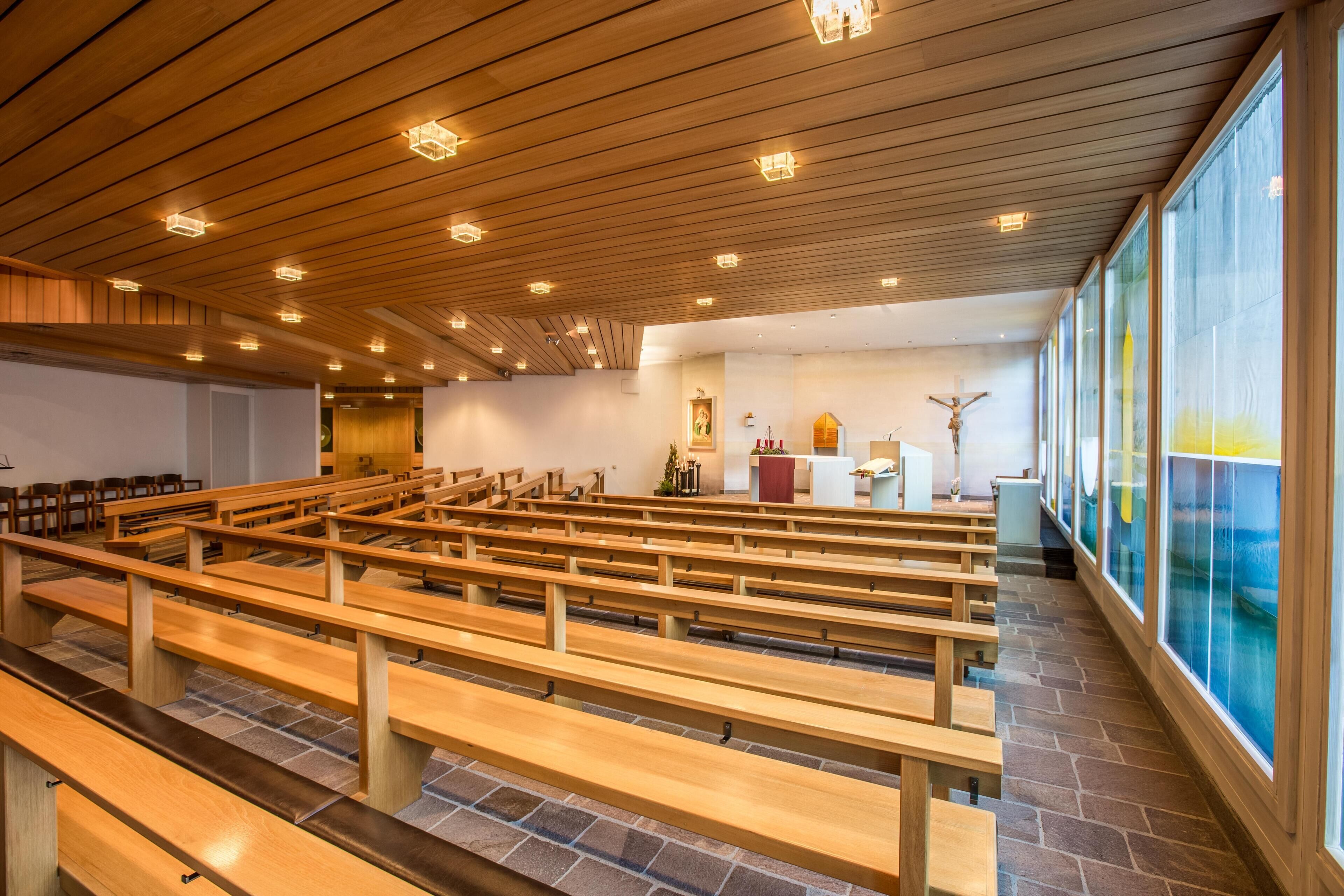 Interior de iglesia con bancos de madera, suelo de piedra y un amplio ventanal con vistas.